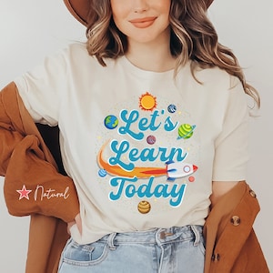 Pode incluir: Uma camiseta branca com um gr&aacute;fico azul e laranja que diz "Let's Learn Today" com planetas e um foguete. A camiseta est&aacute; em uma modelo que usa um chap&eacute;u marrom e jeans azul.