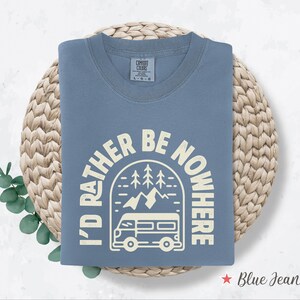 May include: A blue jean colored t-shirt with the text "I'D RATHER BE NOWHERE" in a cream-colored font. The graphic includes a camper van, trees, and mountains. The shirt is folded on a woven mat.