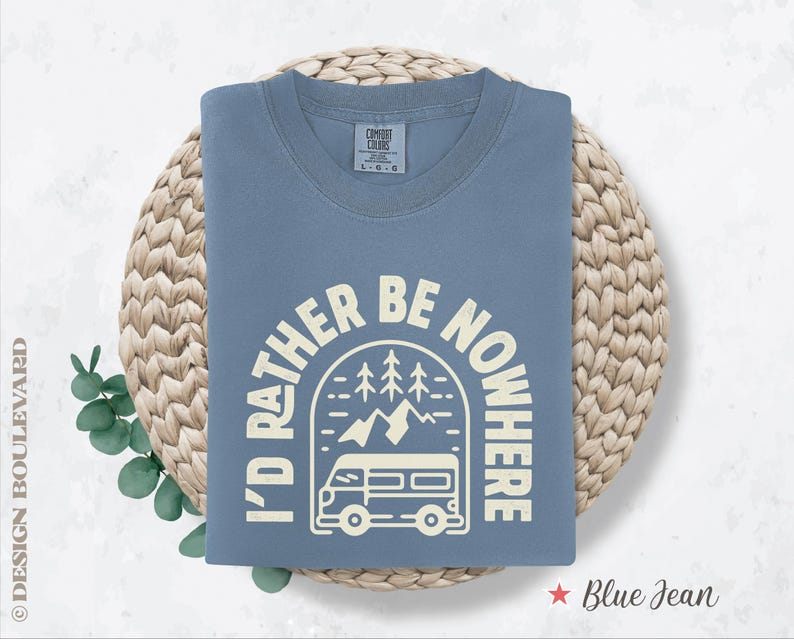 May include: A blue jean colored t-shirt with the text "I'D RATHER BE NOWHERE" in a cream-colored font. The graphic includes a camper van, trees, and mountains. The shirt is folded on a woven mat.