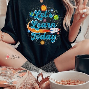 Pode incluir: Camiseta preta com um gr&aacute;fico de planetas, um foguete e o texto "Let's Learn Today".