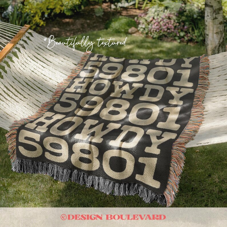 Op de afbeelding: Een zwart en beige geweven plaid met een herhalend patroon van de tekst 59801 en het woord HOWDY in grote blokletters. De deken heeft een getextureerd oppervlak en is afgewerkt met een veelkleurige franjerand aan alle zijden.