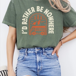 May include: An olive green t-shirt with the text "I'D RATHER BE NOWHERE" in a circular design featuring a camper van, trees, and mountains. The shirt is a comfortable, casual style.
