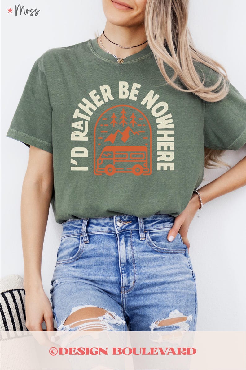 May include: An olive green t-shirt with the text "I'D RATHER BE NOWHERE" in a circular design featuring a camper van, trees, and mountains. The shirt is a comfortable, casual style.