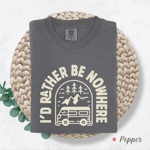May include: A gray t-shirt with the text "I'D RATHER BE NOWHERE" in a cream-colored arched design featuring a camper van, mountains, and trees. The shirt is folded on a woven mat.