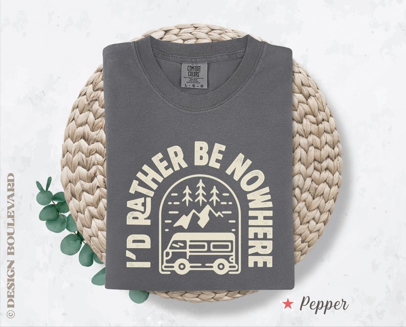 May include: A gray t-shirt with the text "I'D RATHER BE NOWHERE" in a cream-colored arched design featuring a camper van, mountains, and trees. The shirt is folded on a woven mat.