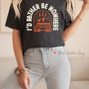 May include: A dark heather grey t-shirt with a graphic of a vintage camper van driving through a mountain landscape. The text "I'd Rather Be Nowhere" is printed in a white arch above the camper van.