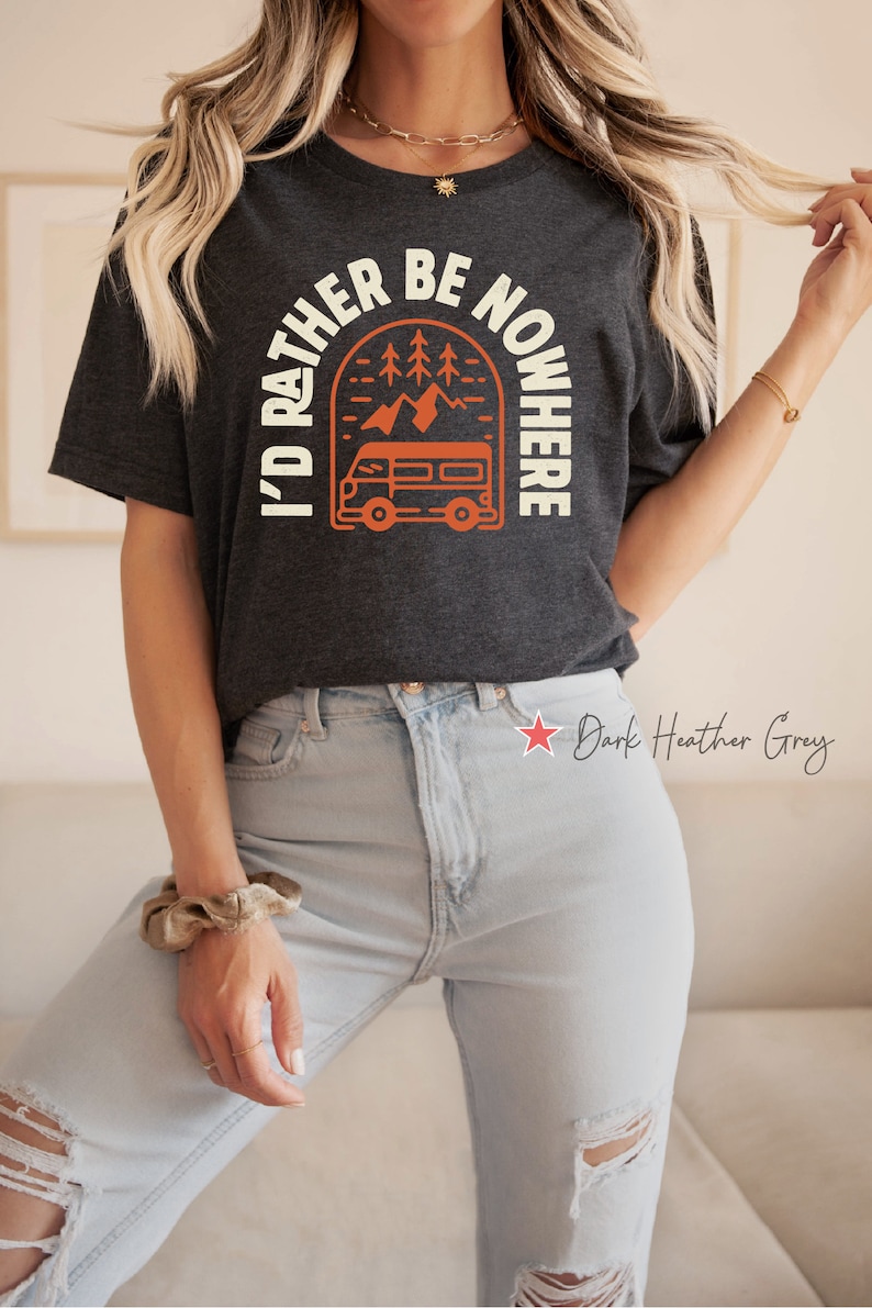 May include: A dark heather grey t-shirt with a graphic of a vintage camper van driving through a mountain landscape. The text "I'd Rather Be Nowhere" is printed in a white arch above the camper van.
