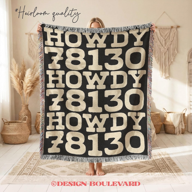 Op de afbeelding: Een zwarte geweven plaid met de herhalende tekst HOWDY 78130 in een groot, cr&egrave;mekleurig bloklettertype. De deken heeft een gefranjerde rand met veelkleurige draden en een getextureerd uiterlijk over het gehele oppervlak van de stof.