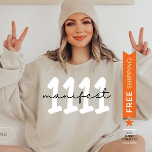 May include: A beige sweatshirt with the text "1111 manifest" in white and black lettering. The sweatshirt is being modeled by a person with long brown hair and a beige beanie.