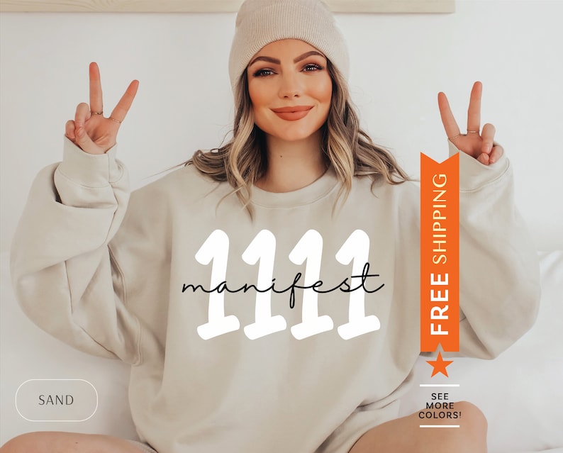 May include: A beige sweatshirt with the text "1111 manifest" in white and black lettering. The sweatshirt is being modeled by a person with long brown hair and a beige beanie.