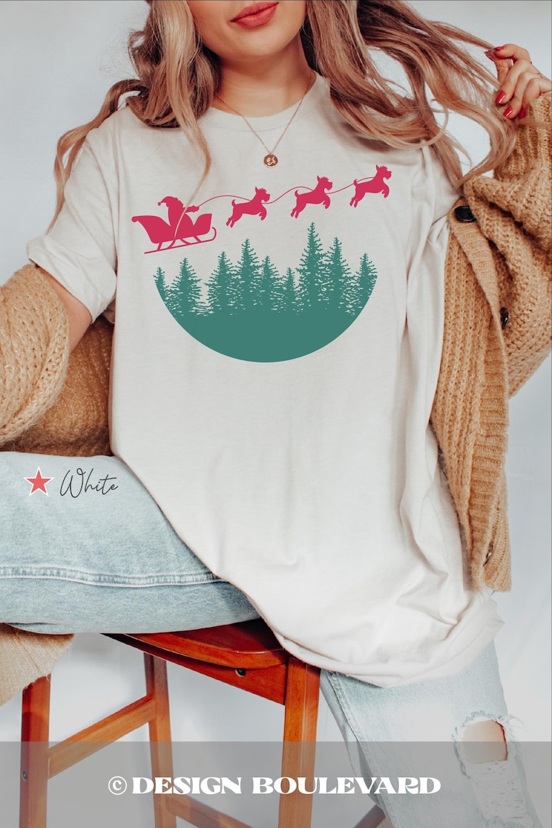 May include: White t-shirt featuring a Santa Claus in a sleigh design pulled by dogs, above a green forest silhouette. The text "White" is displayed on the shirt. The Design Boulevard logo is at the bottom.