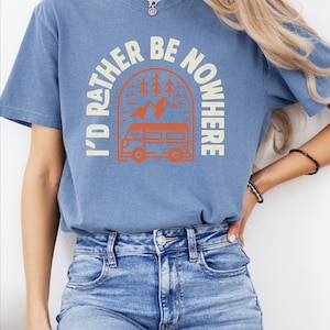 May include: A blue short-sleeved t-shirt with the cream text "I'D RATHER BE NOWHERE". The graphic shows an orange camper van, trees, and mountains. The shirt is worn with blue denim jeans.
