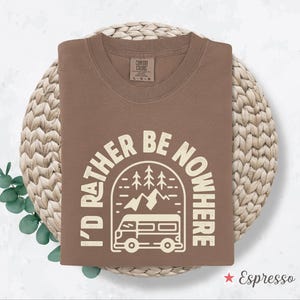 May include: Espresso-colored t-shirt with the text "I'd Rather Be Nowhere" in a white, circular design featuring a camper van, mountains, and trees. The shirt is folded and rests on a woven, circular surface.