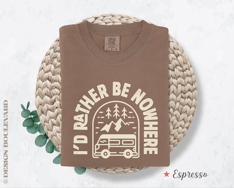 May include: Espresso-colored t-shirt with the text "I'd Rather Be Nowhere" in a white, circular design featuring a camper van, mountains, and trees. The shirt is folded and rests on a woven, circular surface.