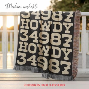 Op de afbeelding: Een zwart en cr&egrave;mekleurige geweven deken met een herhalend patroon van het getal 34987 en het woord HOWDY in een vetgedrukt bloklettertype. De deken heeft een gefranjerde rand en bevat de tekst *Machine washable in de linkerbovenhoek.