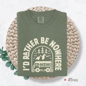 May include: A moss green crew neck sweatshirt with the text "I'D RATHER BE NOWHERE" in a white, circular design featuring a camper van, mountains, and trees. The sweatshirt is folded on a woven, circular mat.