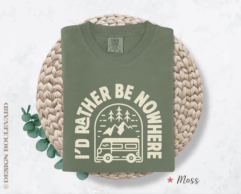 May include: A moss green crew neck sweatshirt with the text "I'D RATHER BE NOWHERE" in a white, circular design featuring a camper van, mountains, and trees. The sweatshirt is folded on a woven, circular mat.