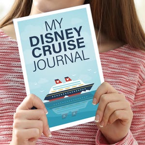 May include: A journal with the text "MY DISNEY CRUISE JOURNAL" on the cover. The cover features a cruise ship illustration on a blue background with clouds and seagulls. The journal is held by a person.