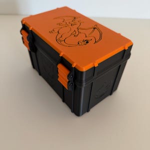 May include: A rectangular storage box with a black base and a bright orange lid. The lid features a black engraved illustration of a winged dragon-like creature. The box has orange side latches and a matching dragon emblem on the black side panel.