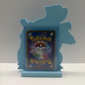 May include: A light blue display stand shaped like a silhouette of a creature, holding a rectangular trading card in a clear protective sleeve. The card features the back design of a Pokémon card with the word Pokémon and a central sphere.