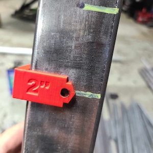 May include: A close-up of a metal beam with a red 2-inch measuring tool attached. The tool is positioned at a marked point on the beam. The text "2" is clearly visible on the tool.