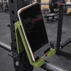 May include: A green phone holder attached to a black weightlifting rack. A smartphone with a white and gold case is placed in the holder. The background shows a gym setting with weights and other equipment.