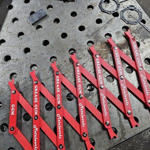 May include: A collection of red grease gun tools with the text "GREASE GUN Metalworks" printed on them. The tools are arranged on a metal surface with other metal components and tools.