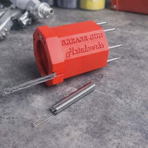 May include: A bright red, hexagonal-shaped grease gun component with the embossed text "GREASE GUN Metalworks" on its side. It features four protruding metal prongs, a flat metal lever attached to the front, and a separate silver metal coil spring.