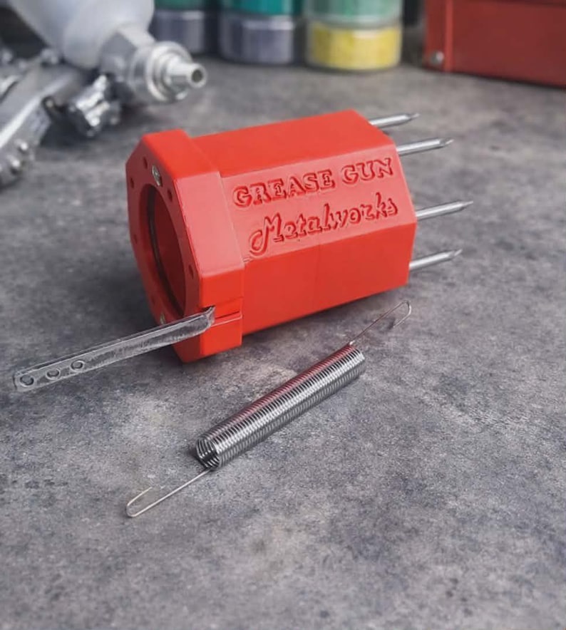 May include: A bright red, hexagonal-shaped grease gun component with the embossed text "GREASE GUN Metalworks" on its side. It features four protruding metal prongs, a flat metal lever attached to the front, and a separate silver metal coil spring.