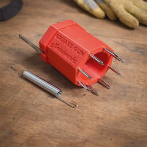 May include: A bright orange, hexagonal-shaped grease gun tool with the embossed text "GREASE GUN Metalworks" on its side. The tool features six sharp, silver-colored metal needles protruding from one end and a separate silver metal tension spring.