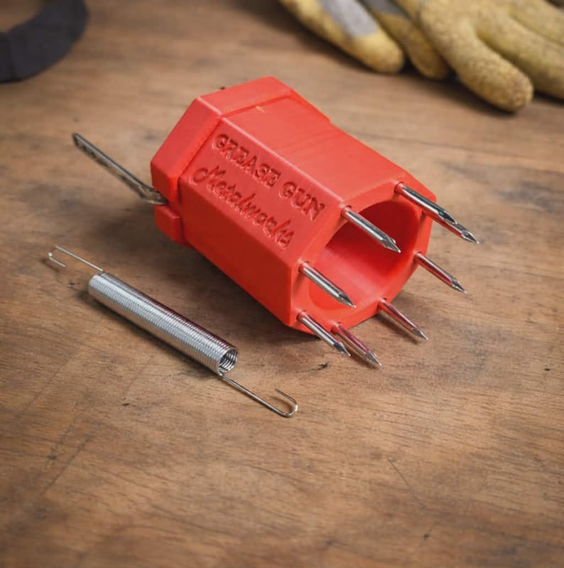 May include: A bright orange, hexagonal-shaped grease gun tool with the embossed text "GREASE GUN Metalworks" on its side. The tool features six sharp, silver-colored metal needles protruding from one end and a separate silver metal tension spring.