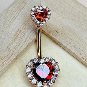 May include: A gold belly button ring with two heart-shaped charms. Each charm features a red gemstone surrounded by a halo of clear cubic zirconia stones.
