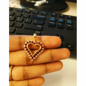 Peut inclure: Un pendentif en or en forme de cœur avec des pierres précieuses rouges et blanches. Le pendentif est sur une main.