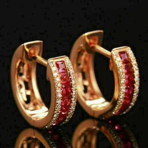 May include: A pair of gold hoop earrings with a row of red and white gemstones. The earrings are made of gold and have a polished finish.