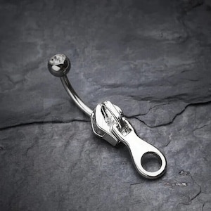 Puede incluir: Piercing corporal de plata con una gema transparente y un dije en forma de cremallera.