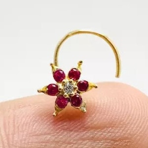 May include: Gold nose ring featuring a red and white flower design. The flower has a single white diamond in the centre and red gemstones around it.