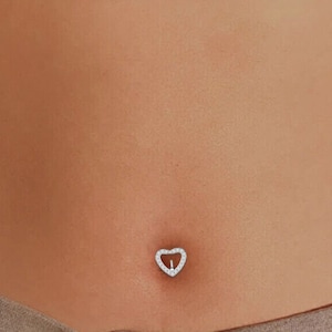 Könnte beinhalten: Ein silberner Bauchnabelring mit einem herzförmigen Design. Das Herz ist mit kleinen, klaren Steinen verziert.