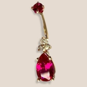 Pode incluir: Um anel de umbigo de ouro com uma gema de rubi em forma de lágrima e uma gema de rubi menor na parte superior. O rubi em forma de lágrima é cercado por gemas menores e transparentes.