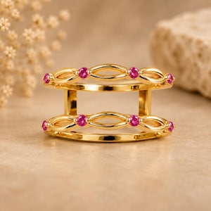 May include: A double-band gold ring featuring two parallel bands connected by vertical gold bars. Each band is designed with an open, repeating oval pattern, and each oval is accented with a small, round, vibrant pink gemstone at its center.