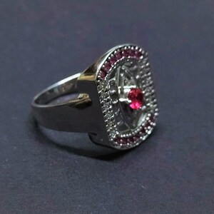 Könnte beinhalten: Ein silberner Ring mit einem roten Edelstein, der von kleineren weißen Edelsteinen und einem rosa Steinrand umgeben ist.