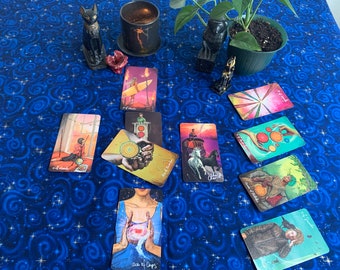 May include: A tarot card reading spread on a blue tablecloth with a star pattern. The cards are spread out in a circle and show various images, including a person riding a horse, a person holding a cup, and a person holding a sword.
