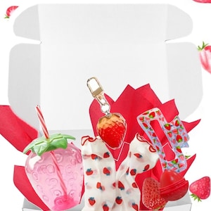 May include: A white gift box with a red tissue paper lining. The box contains a pink strawberry-shaped drink cup with a straw, a pair of white socks with strawberry prints, a clear plastic strawberry-shaped keychain, and a red strawberry-shaped lip balm.