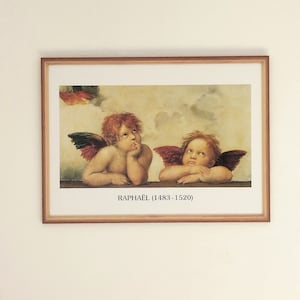 Puede incluir: Una impresión enmarcada de una pintura renacentista que representa a dos querubines con alas rojas, uno con un dedo en los labios y el otro mirando hacia arriba. La pintura se atribuye a Rafael (1483-1520).