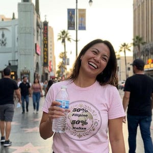 Puede incluir: Una mujer sonríe, vistiendo una camiseta rosa claro con el texto "50% Filipino 50% Italian", sosteniendo una botella de agua. El fondo muestra una calle con gente y edificios, incluido el Teatro El Capitán.