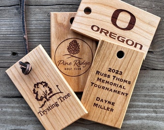 May include: Four wooden plaques with engraved text and logos. The top plaque reads "O OREGON". The plaque below it reads "Pine Ridge Golf Club" with a pine tree logo. The plaque to the right reads "2023 Russ Thoms Memorial Tournament Dayne Miller". The bottom plaque reads "Trysting Tree" with a tree logo.