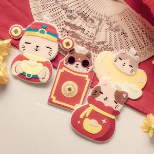 Puede incluir: Una colección de adornos de fieltro con forma de gato en rojo, amarillo y marrón. Un gato viste un atuendo tradicional chino, mientras que otros sostienen sobres rojos y lingotes de oro. Los adornos están decorados con pequeños detalles y sobre un fondo rojo.