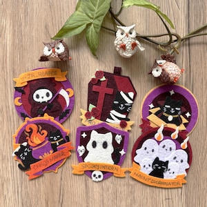 May include: A collection of felt patches and three small owl figurines. The patches feature designs including a grim reaper, a mummy in a coffin, a black cat with a cauldron, a ghost, and a group of ghosts, with text reading JR. REAPER, CHAOS MAKER, SPOOKY INTERN, and HAUNT COORDINATOR.