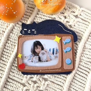 Puede incluir: Un marco de fotos de fieltro con forma de televisor retro, coronado por un gato azul oscuro. El marco contiene una foto de una persona con dos gatos. Está decorado con estrellas amarillas, peces azules y un corazón rojo.