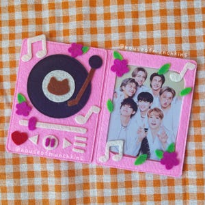 May include: A pink felt photo album with a music theme. The left side features a record player design with a cat-shaped detail. The right side holds a photo of a group of people. Decorative musical notes and flowers surround the photo.