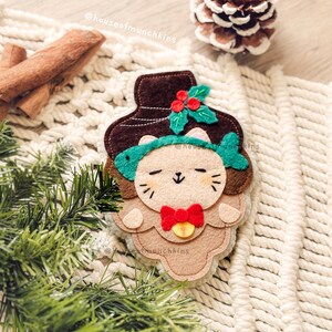Puede incluir: Adorno de gato de fieltro hecho a mano con sombrero marrón, diadema de pez verde y pajarita roja. El gato tiene los ojos cerrados y una pequeña campana. El adorno está decorado con acebo y bayas, ideal para Navidad.